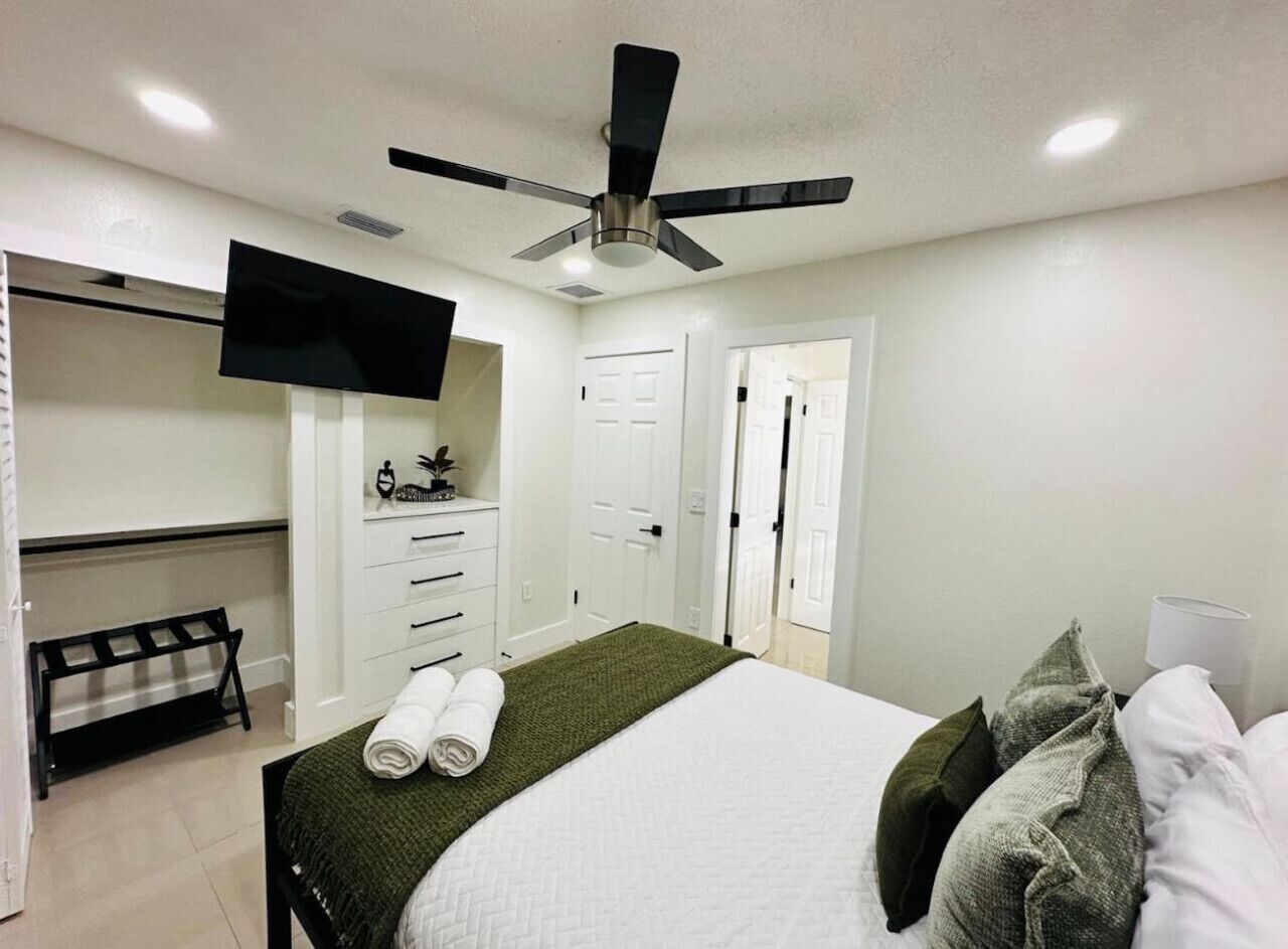 Une chambre à coucher avec un lit, un ventilateur de plafond, un téléviseur fixé au mur et un banc.