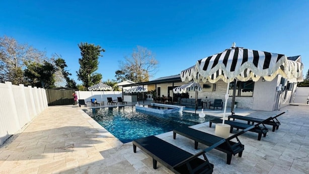 A swimming pool with lounge chairs and umbrellas, surrounded by a white fence and trees. - Pool+Spa+ 3.5 Baths+Outdoor Grill (Bradenton)