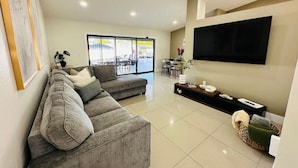 A modern living room with a grey sectional sofa, a flat-screen TV mounted on the wall, and a large window offering a view of the outdoors. - Pool+Spa+ 3.5 Baths+Outdoor Grill (Bradenton)