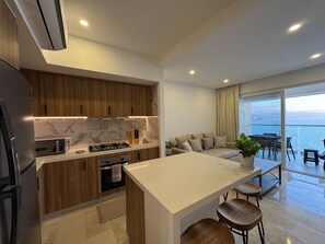 A modern kitchen with wooden cabinets, a white countertop, and a built-in oven. There is a dining area with a table and chairs, and a large window offering a view of the sea. - Ocean view loft, Harbor 171 (Puerto Vallarta)