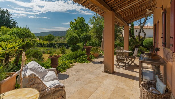Un patio cubierto con mobiliario exterior, una mesa con una parrilla y vistas a un jardín y colinas.