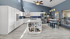 Une cuisine moderne avec des meubles blancs, un îlot central orné de bouteilles, un ventilateur de plafond et un coin repas avec une table et des chaises.