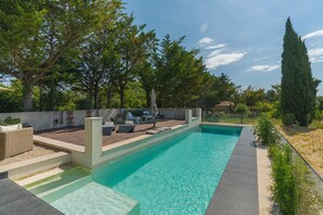A rectangular swimming pool with a wooden deck, lounge chairs, and a tall tree in the background.