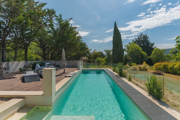 A rectangular swimming pool with a wooden deck and lounge chairs, surrounded by greenery and a tall tree.