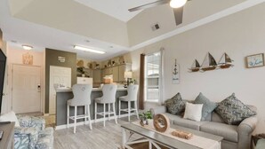 A spacious living area with a grey sofa, a coffee table, and a kitchen area with bar stools. - The Fairways at River Oaks Retreat (Myrtle Beach)