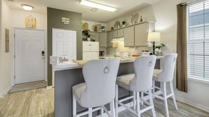 A modern kitchen with a central island, bar stools, and a window with blinds. - The Fairways at River Oaks Retreat (Myrtle Beach)