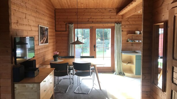 Interior d'una cabana de fusta amb una taula de menjador, cadires, un televisor i una zona de cuina.