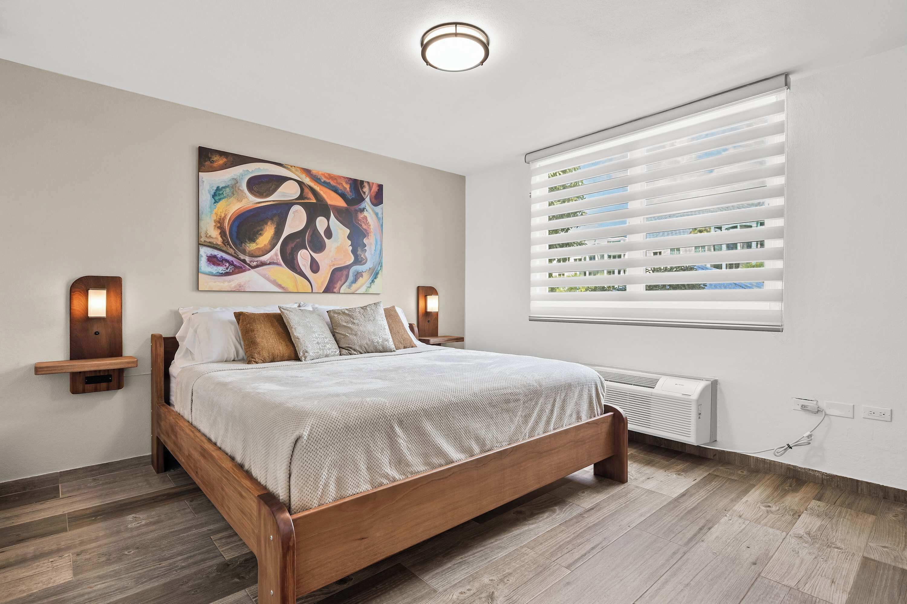 Une chambre avec un lit en bois, un grand tableau abstrait, une fenêtre avec des stores et un radiateur fixé au mur.