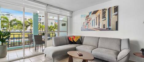 A modern living room with a grey sofa, a small coffee table, and a large window offering a view of a pool and buildings.