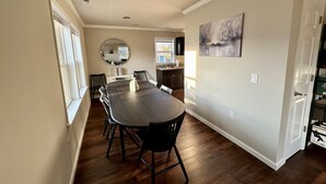 Een eetgedeelte met een grote houten tafel en stoelen, een keuken op de achtergrond, en een schilderij aan de muur.