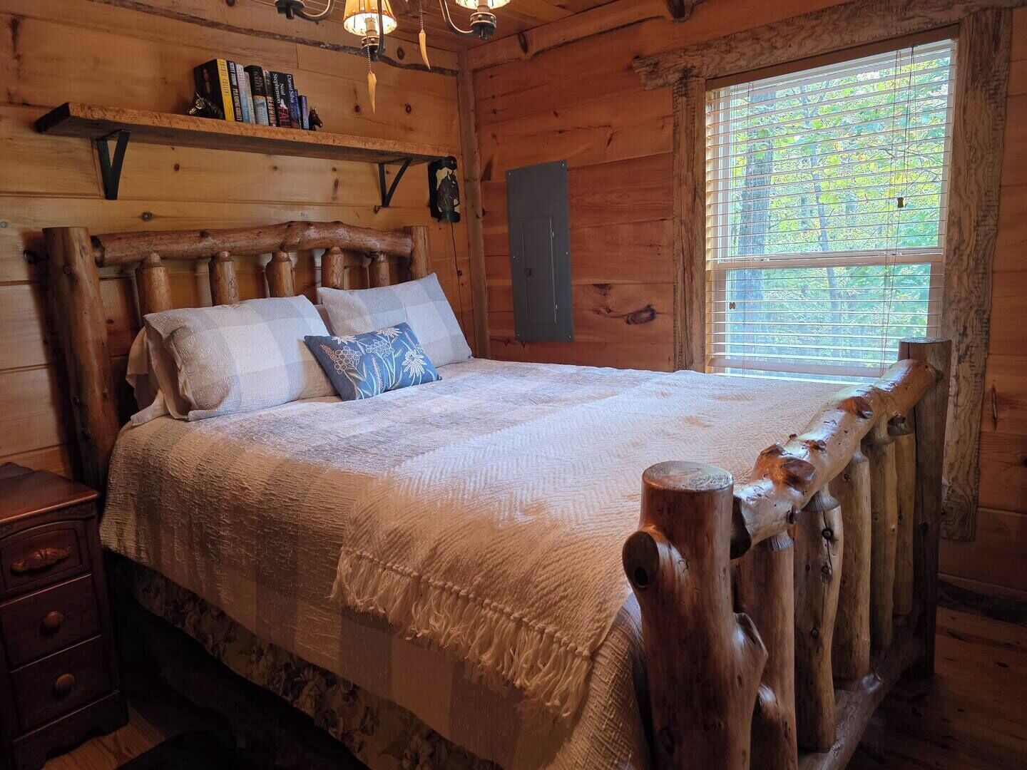 A wooden cabin bedroom with a bed, a nightstand, a shelf with books, and a window with blinds.