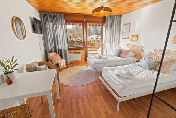 A modern bedroom with two single beds, a small table, a chair, a potted plant, and a view of the outdoors.