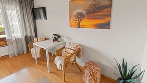 Una habitación con una mesa blanca, sillas de mimbre, un otomán estampado con motivos de cebra, una planta en maceta y un cuadro de un árbol y una puesta de sol en la pared.