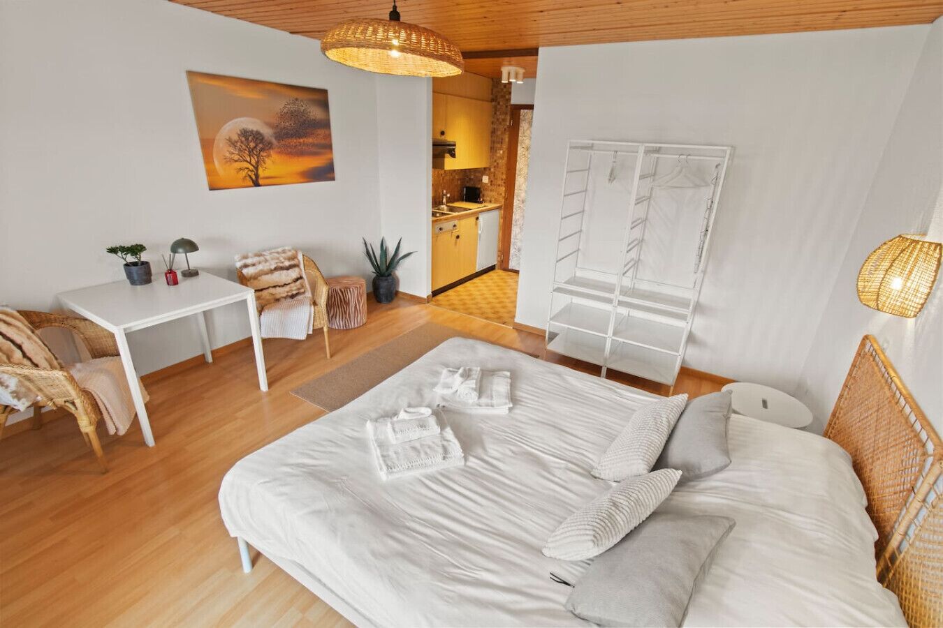 Une chambre moderne avec un grand lit, un bureau et une chaise. On aperçoit une cuisine au fond.
