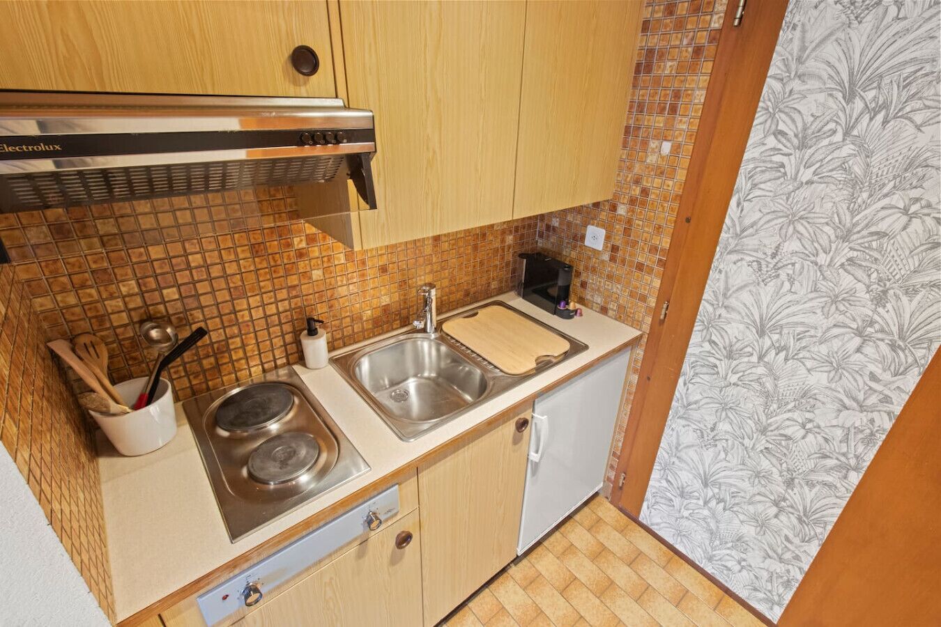 Une cuisine de taille réduite, équipée d’une cuisinière à gaz, d’un évier à deux bacs et de meubles de rangement en bois.