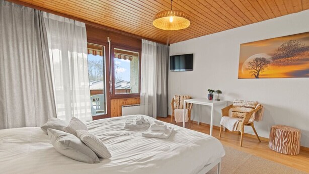 Una cama bien hecha con almohadas y una toalla doblada, una mesita con una silla, un televisor en la pared y un cuadro de un árbol y un atardecer.