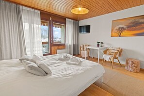A neatly made bed with pillows and a folded towel, a small table with a chair, a television on the wall, and a painting of a tree and sunset. - Charmant studio avec balcon à 5 min du lac (Portalban)