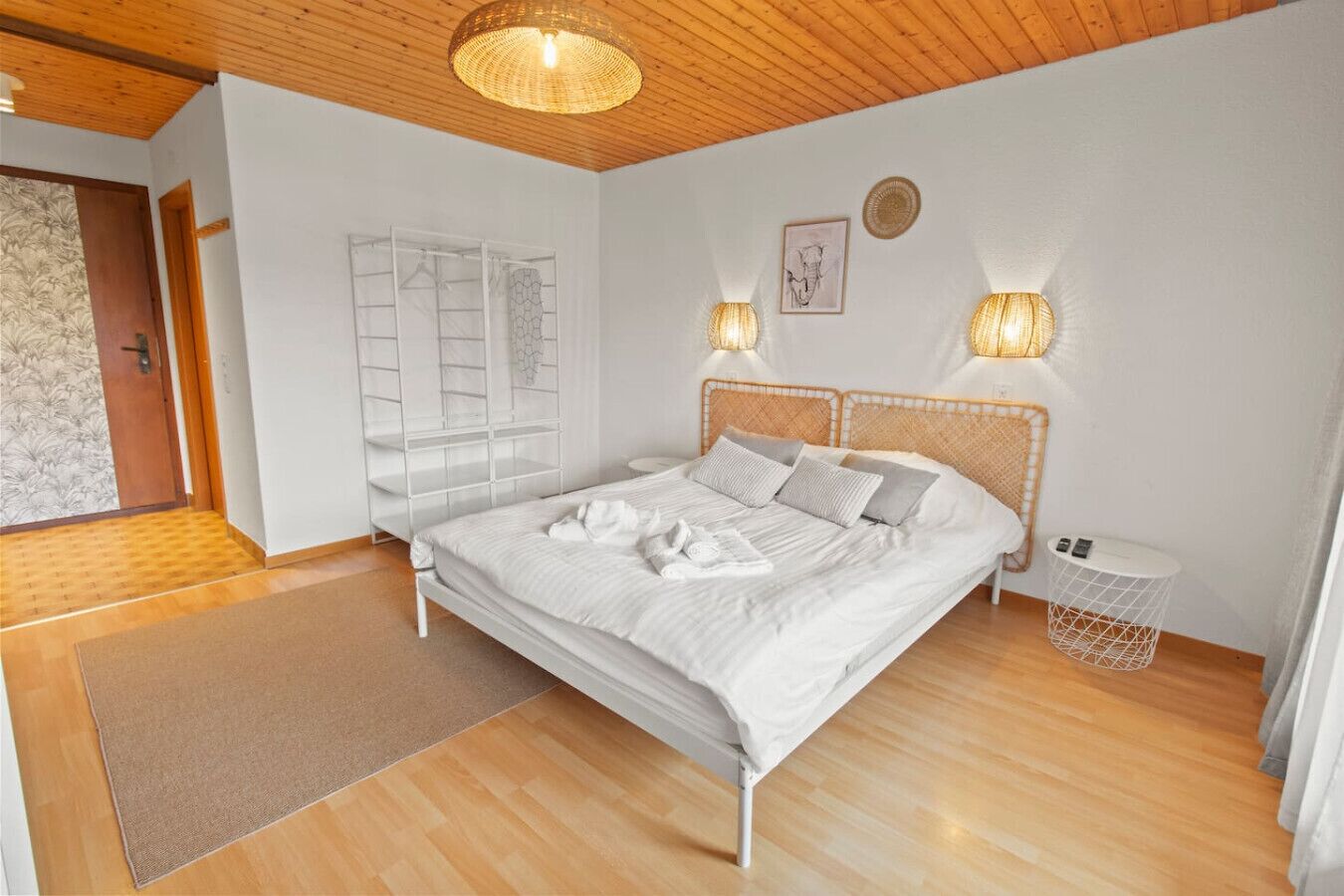 A bedroom with a wooden floor, a white bed with grey pillows, two wall-mounted lamps, a small white side table, and a wicker basket.