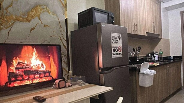 A kitchen with a marble backsplash, a television displaying a fireplace, a refrigerator, and a dining area with a chair. - Misty Dream Condostay (Pasay City)