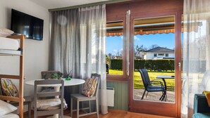 Une pièce avec un plancher en bois, une table ronde entourée de chaises, un téléviseur fixé au mur et une porte-fenêtre coulissante donnant sur un espace extérieur aménagé avec des chaises.