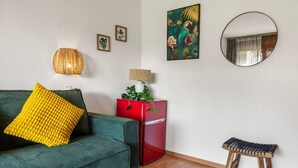Un salon avec un canapé vert, un coussin jaune, un meuble rouge, un miroir rond et un petit tabouret.