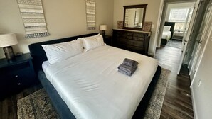 A neatly made bed with white linens, a dark headboard, and a folded towel on the foot of the bed. There is a nightstand with a lamp and a wall hanging above the bed. - Summit Vista Lodge - Breathtaking 180°+ Mountain Views | Hot Tub (Waynesville)