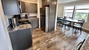 A modern kitchen with dark cabinets, stainless steel appliances, and a dining area with a large window. - Summit Vista Lodge - Breathtaking 180°+ Mountain Views | Hot Tub (Waynesville)