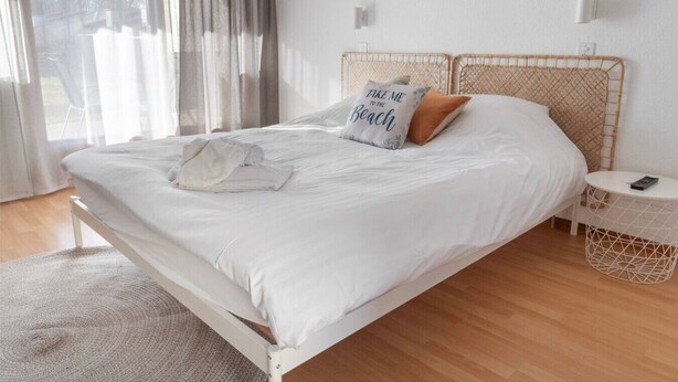 Un lit bien fait, avec des draps blancs, un oreiller orné d’un motif et une serviette pliée sur un plancher en bois.