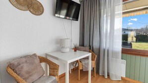 Une pièce avec une table blanche, deux chaises en osier, un téléviseur fixé au mur et une vue sur l’extérieur par une fenêtre ornée de rideaux.