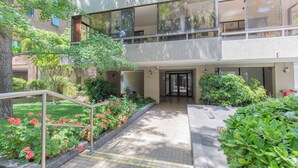 Un edificio moderno con un acceso de doble altura, un pequeño jardín con flores rojas y una barandilla de metal.