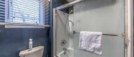 A bathroom with a glass-enclosed shower, a toilet, and a window with blinds.