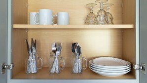 A wooden cabinet with two shelves; the top shelf holds white mugs and wine glasses, and the bottom shelf contains silverware in glass jars and a stack of white plates. - Studio South (On UT Campus & Free Parking) (Knoxville)