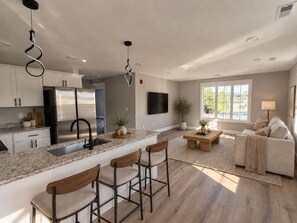 A modern kitchen with a stainless steel refrigerator, a bar area with stools, a dining table with chairs, and a living area with a sofa and a TV.