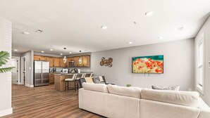 Una cocina moderna con armarios de madera y electrodomésticos de acero inoxidable, un comedor con mesa y sillas blancas, y una sala de estar con sofá color beige y un cuadro colorido en la pared.