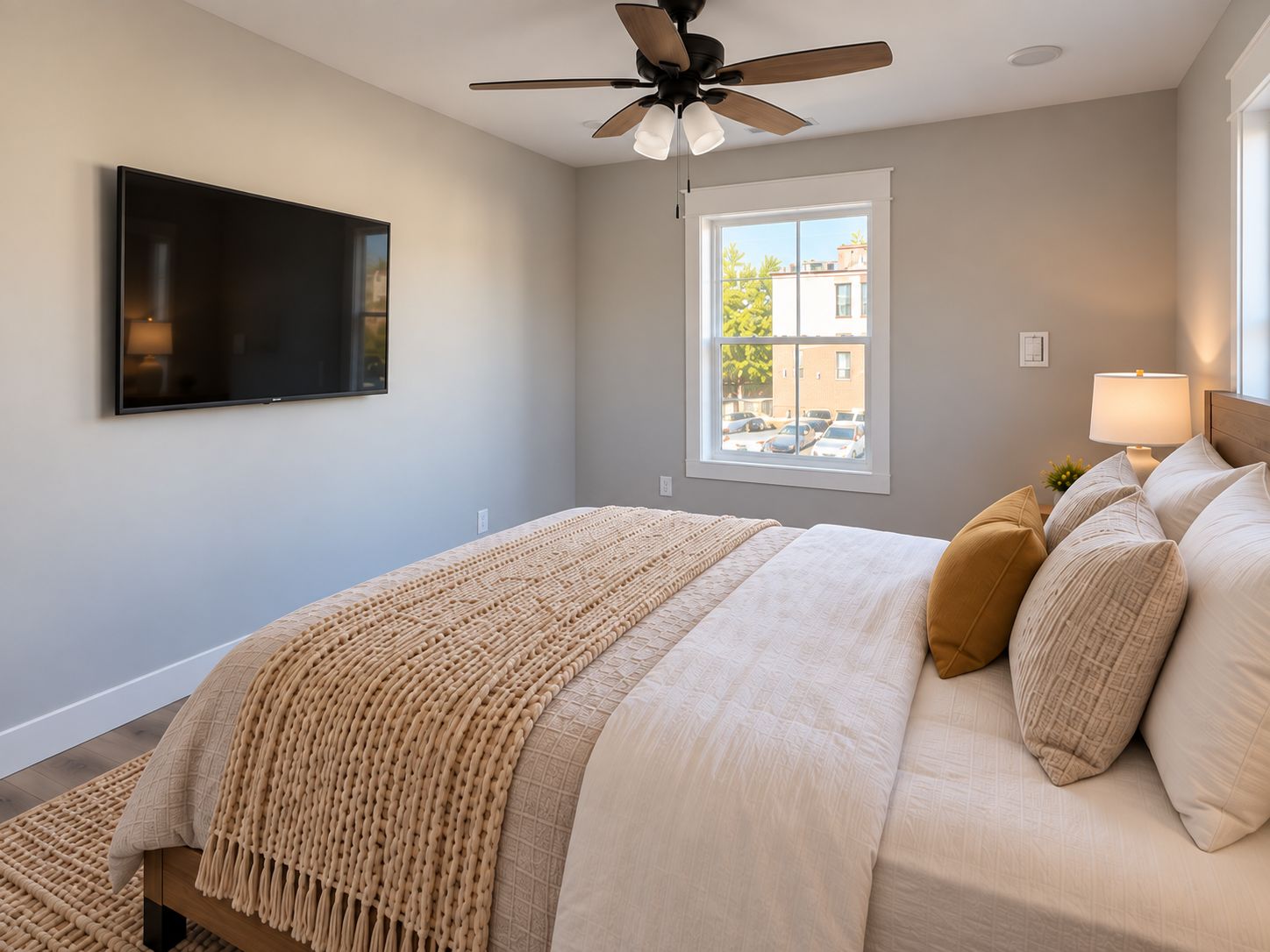 A bedroom with a bed, a ceiling fan, a TV mounted on the wall, and a window with a view of buildings and cars.