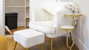 Ein weißer Sessel mit Fußhocker, ein kleiner runder Tisch mit goldfarbener Basis und eine weiße Decke auf dem Sessel.
