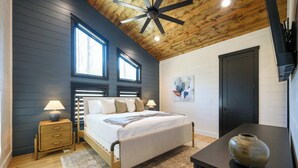 Quarto com cama, mesas de cabeceira, ventoinha no teto, janela e porta.