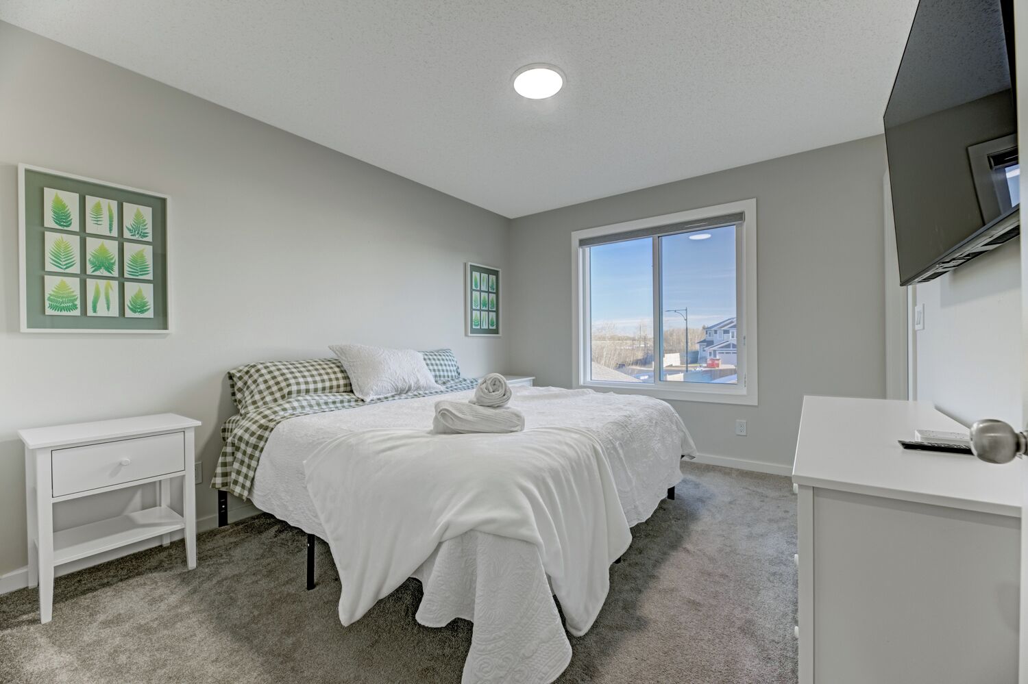 A bedroom with a bed, a bedside table, a dresser, a window, and a wall-mounted TV.