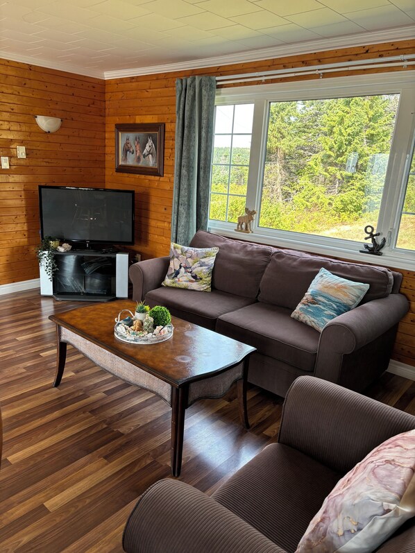 A cozy living room with a wooden floor, a brown sofa, a coffee table with decorative items, a television, and a window with a view of greenery.