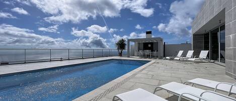 A rooftop pool area with a clear view of the ocean, a covered seating area, and lounge chairs.