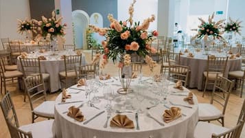 Un salón de banquetes con mesas redondas dispuestas para un evento formal, cada una con un centro de flores y servilletas dobladas con cuidado.