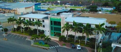 A hotel with a prominent green and white facade, surrounded by parking and palm trees.