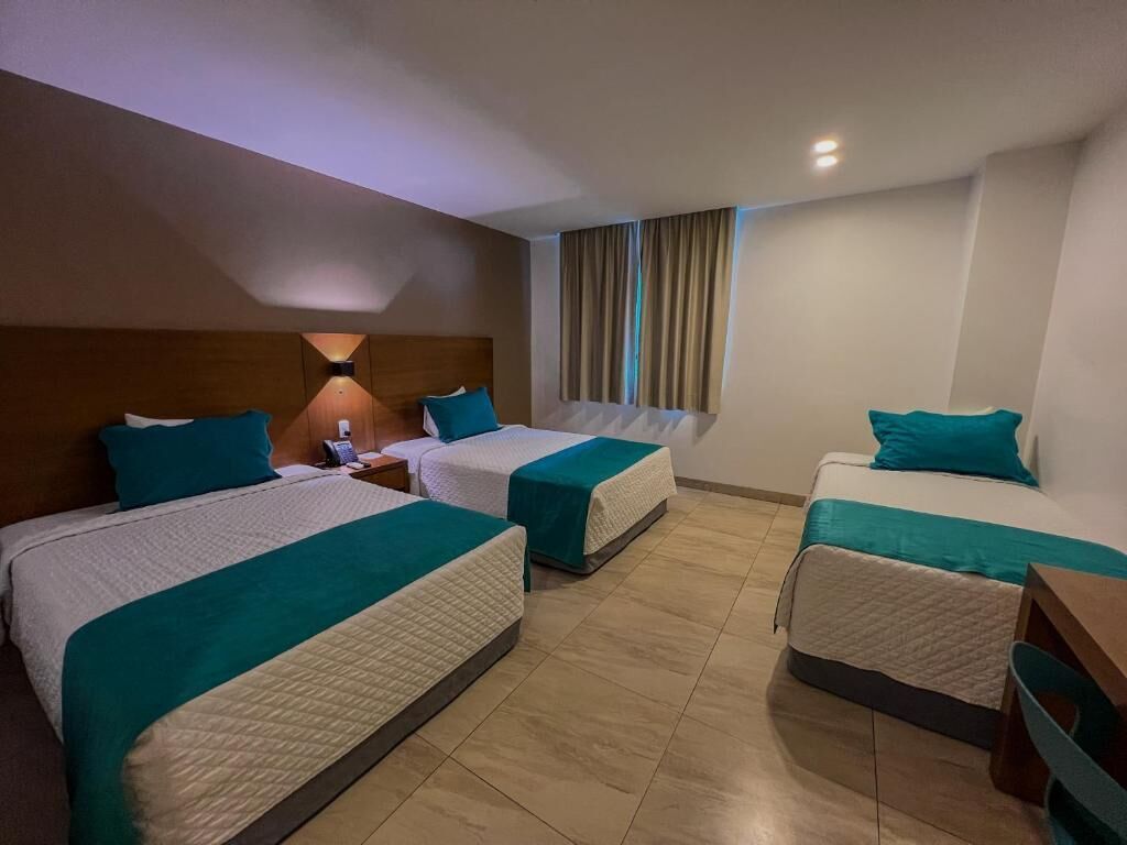 A hotel room with three beds, each with teal bedding and a wooden headboard. There is a nightstand with a lamp and a phone on one side. The room has a tiled floor and a window with curtains.