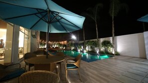 Un espace extérieur avec une piscine, une table et des chaises, entouré de palmiers et éclairé par un éclairage d’ambiance.