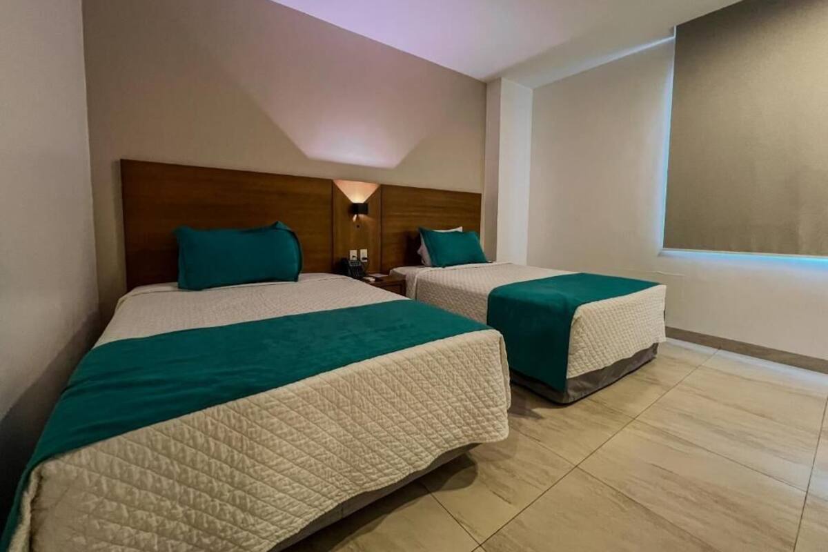 Deux lits doubles avec une literie de couleur turquoise et une tête de lit en bois, une table de chevet avec une lampe, et une grande fenêtre donnant sur un beau paysage.