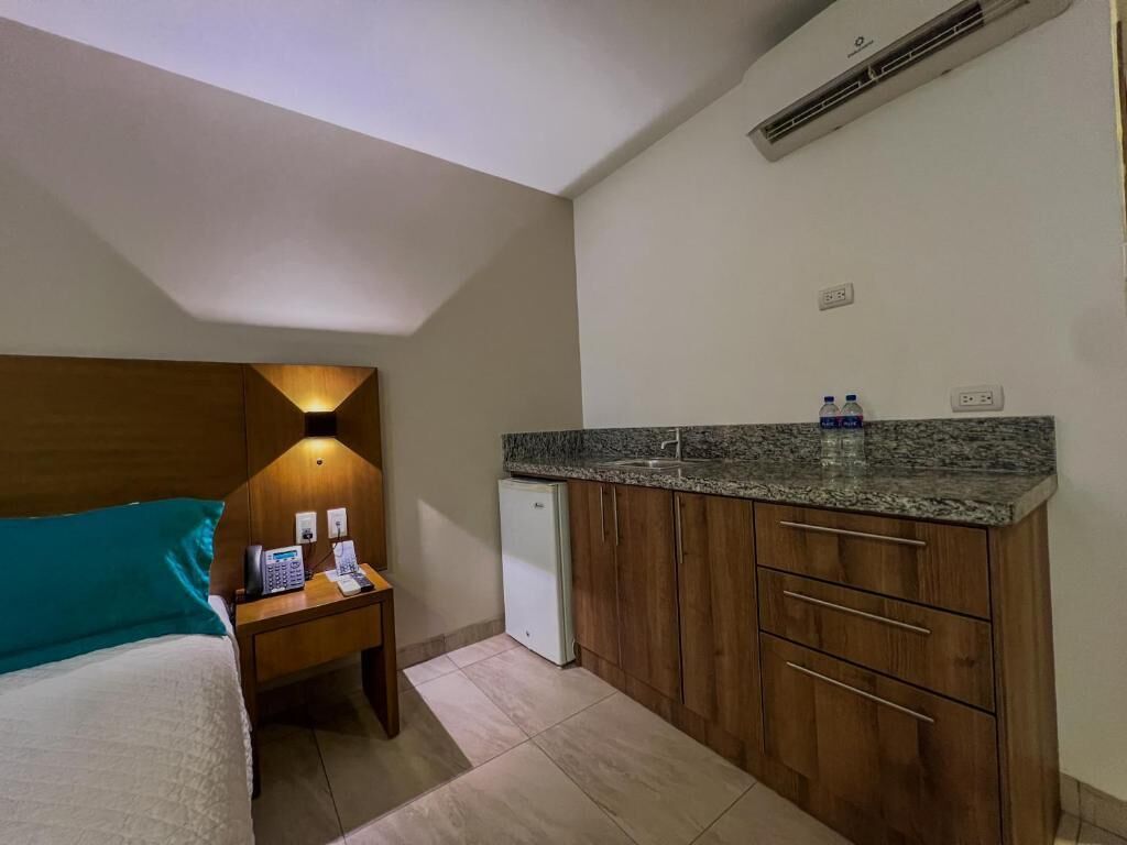 Et kompakt hotelværelse med en seng, et natbord med telefon og lampe, et lille tekøkken med et køleskab og en vanddispenser.