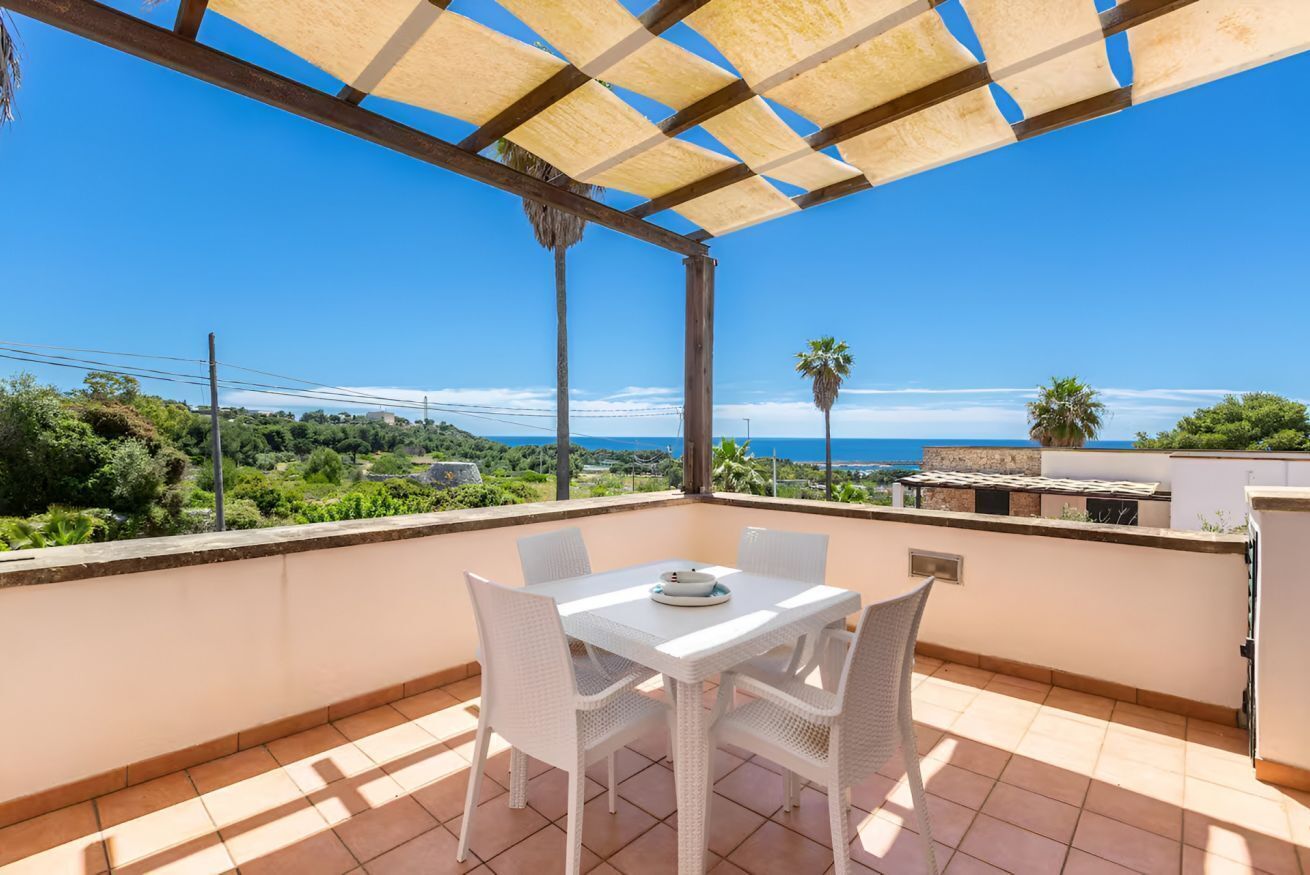 Un balcon avec une table et des chaises blanches, une pergola et une vue sur la mer et la verdure.