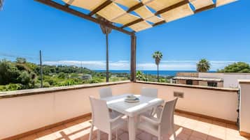 Un balcon avec une table et des chaises blanches, une pergola et une vue sur la mer et la verdure.