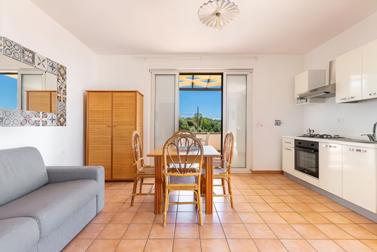 Une cuisine moderne avec des meubles blancs, une table à manger avec des chaises, un canapé gris et une vue à travers une porte vitrée.