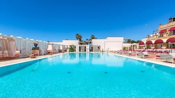 Une piscine d’un bleu éclatant, entourée de petits pavillons blancs et de parasols rouges, avec des bâtiments en arrière-plan.